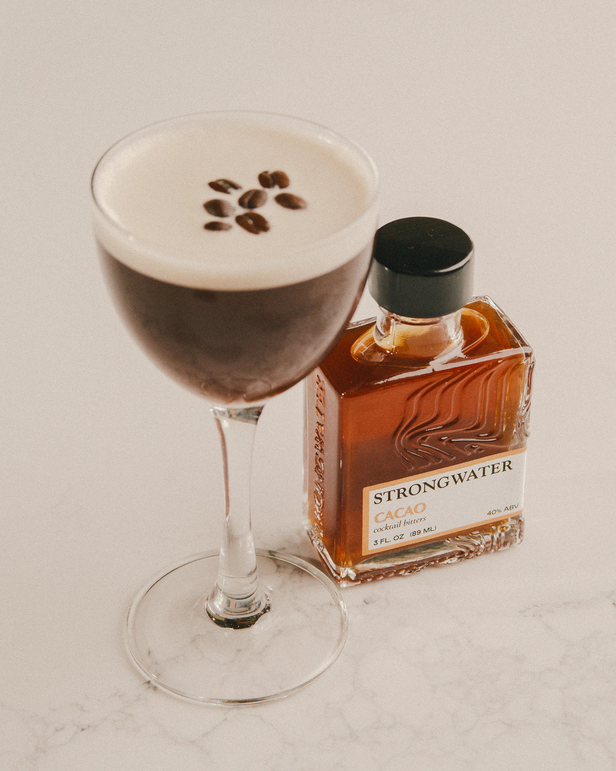 cocktail with coffee beans and a bottle of strongwater cacao bitters on a light background
