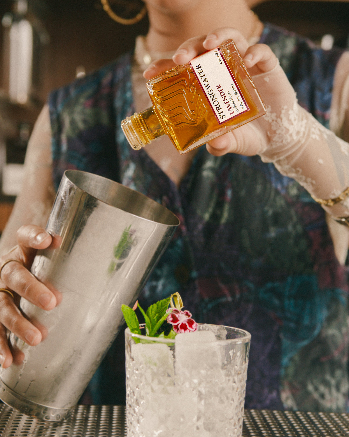 person dashing a bottle of strongwater lavender bitters into a shaker above a glass with ice and garnish.