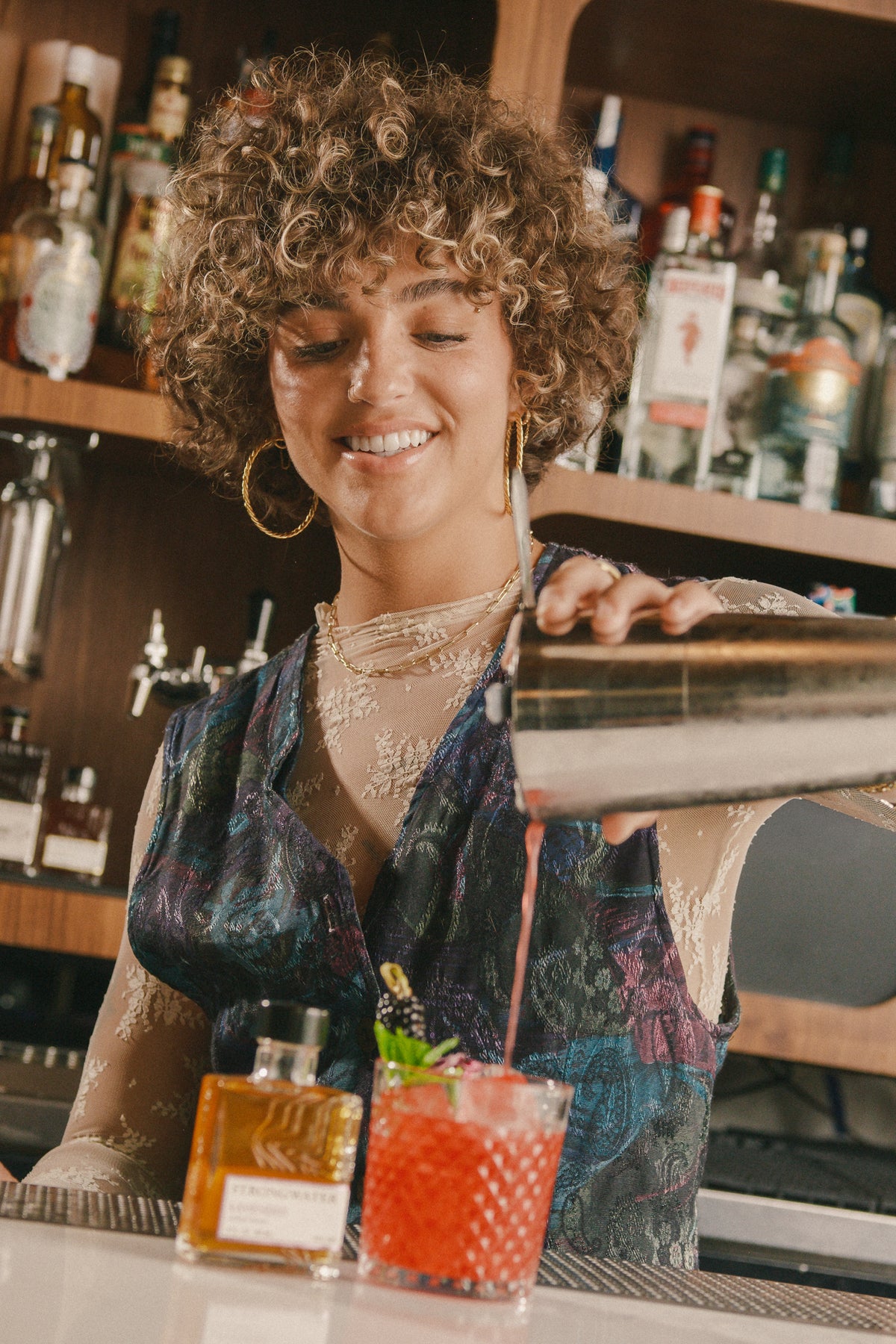 woman pouring a cocktail at a bar with a shaker and a bottle of strongwater lavender bitters