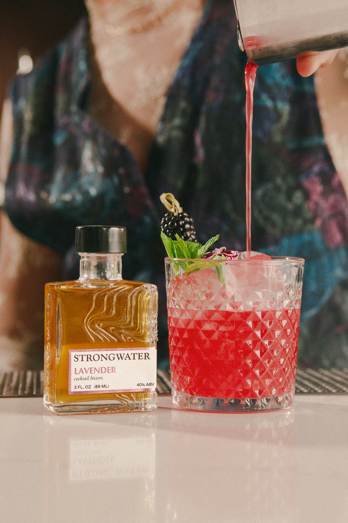 bottle of strongwater lavender bitters next to a cocktail with a red drink and blackberries being poured.