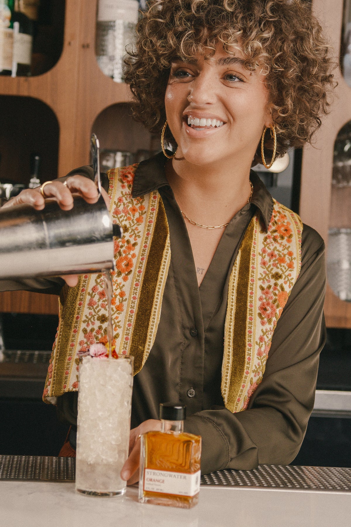 woman in a bar setting with a cocktail shaker and bottle of strongwater orange bitters.