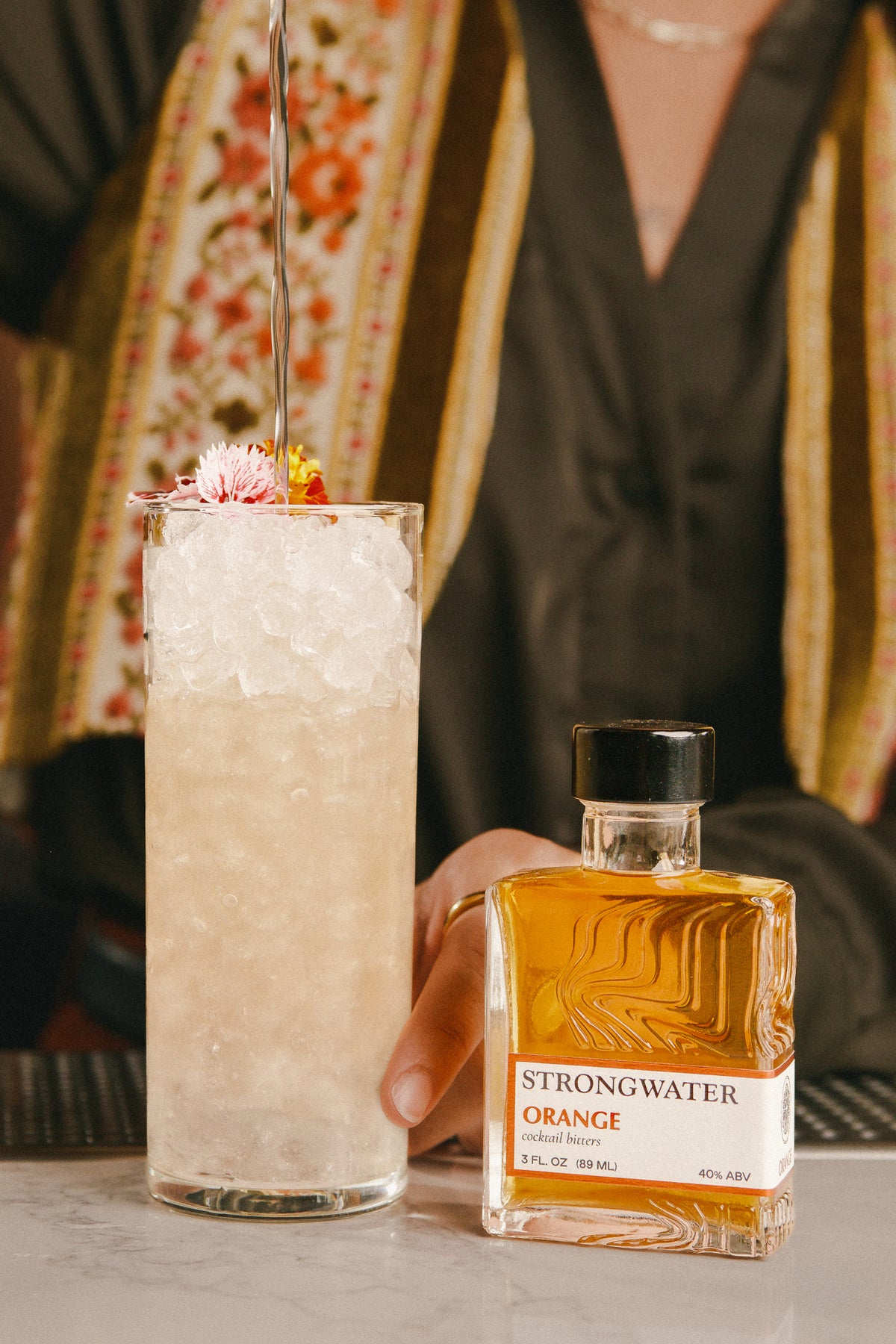 cocktail in a tall glass with ice and a bottle of strongwater orange bitters on a bar counter.